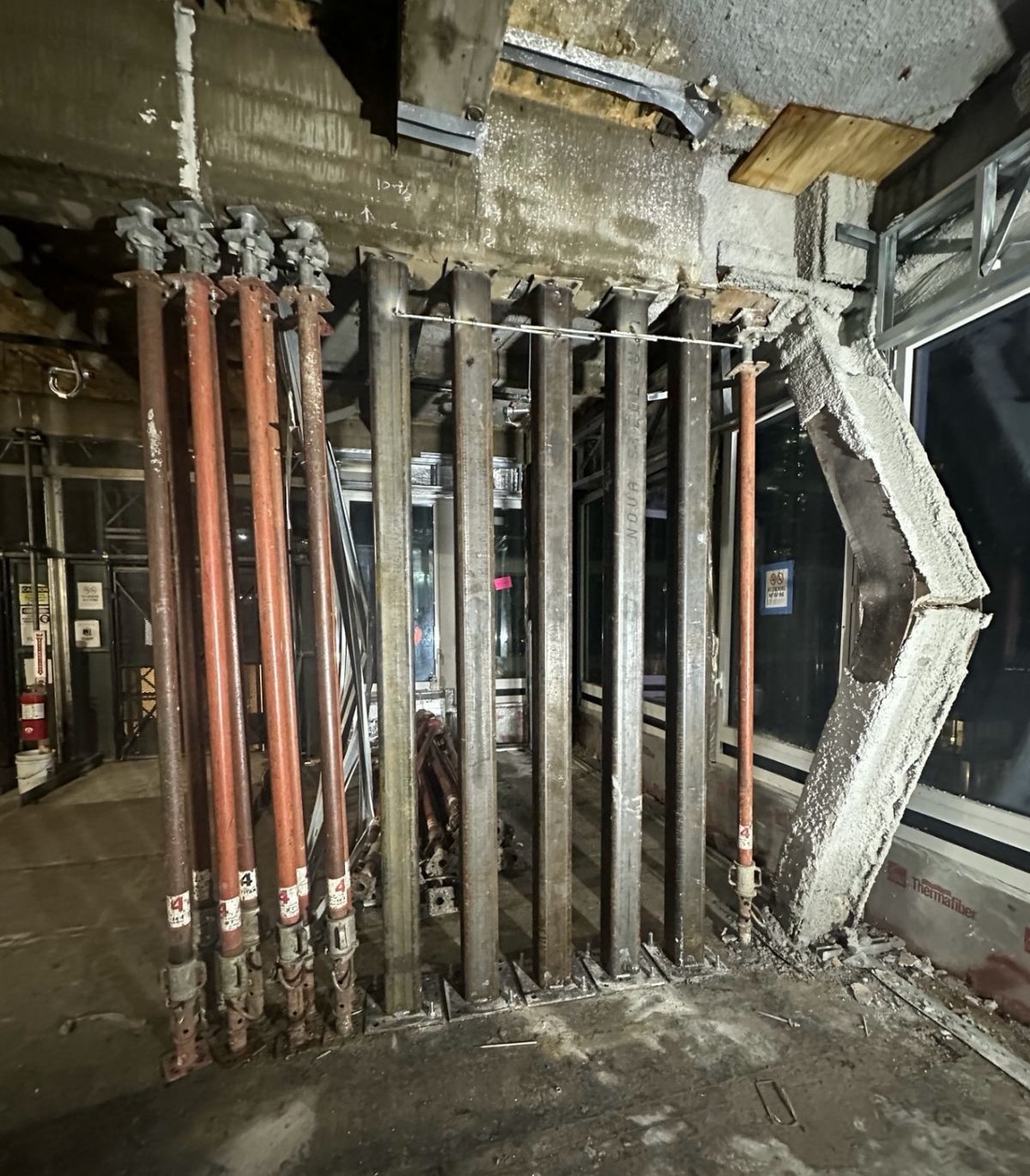 大樓室內施工現場，一整排臨時支撐鋼柱頂住天花板，右側牆面混凝土剝落，露出結構